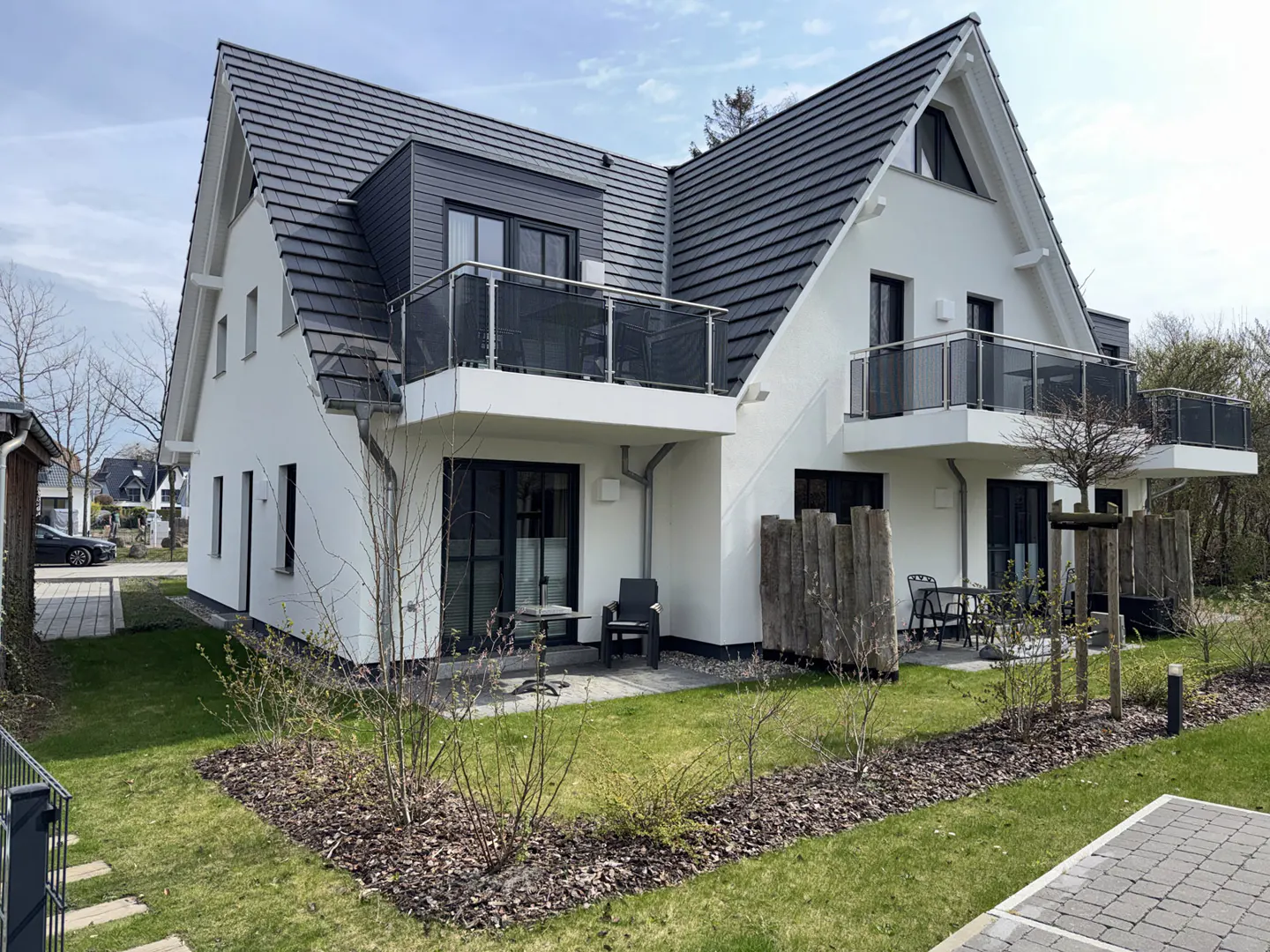 Two-story white house with black trim and a dark gray roof. Balconies with glass railings. Patio with outdoor furniture. Green lawn and landscaping.