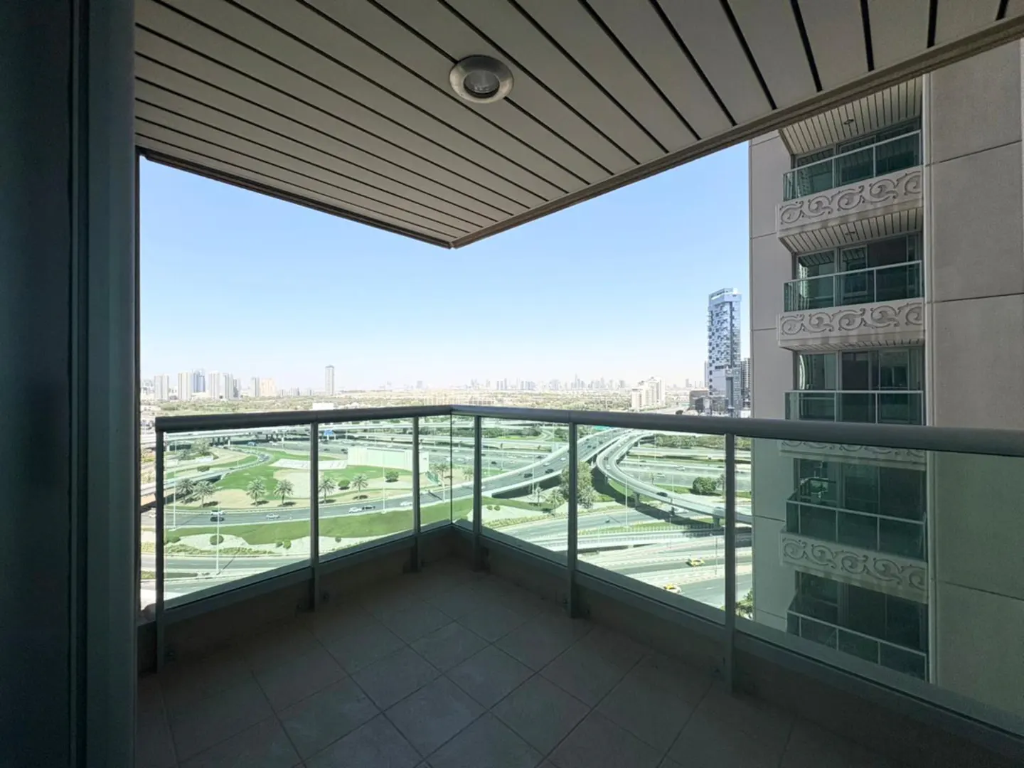 Balcony view of a city skyline. Glass railings surround the tiled balcony. A golf course and highway are visible below.