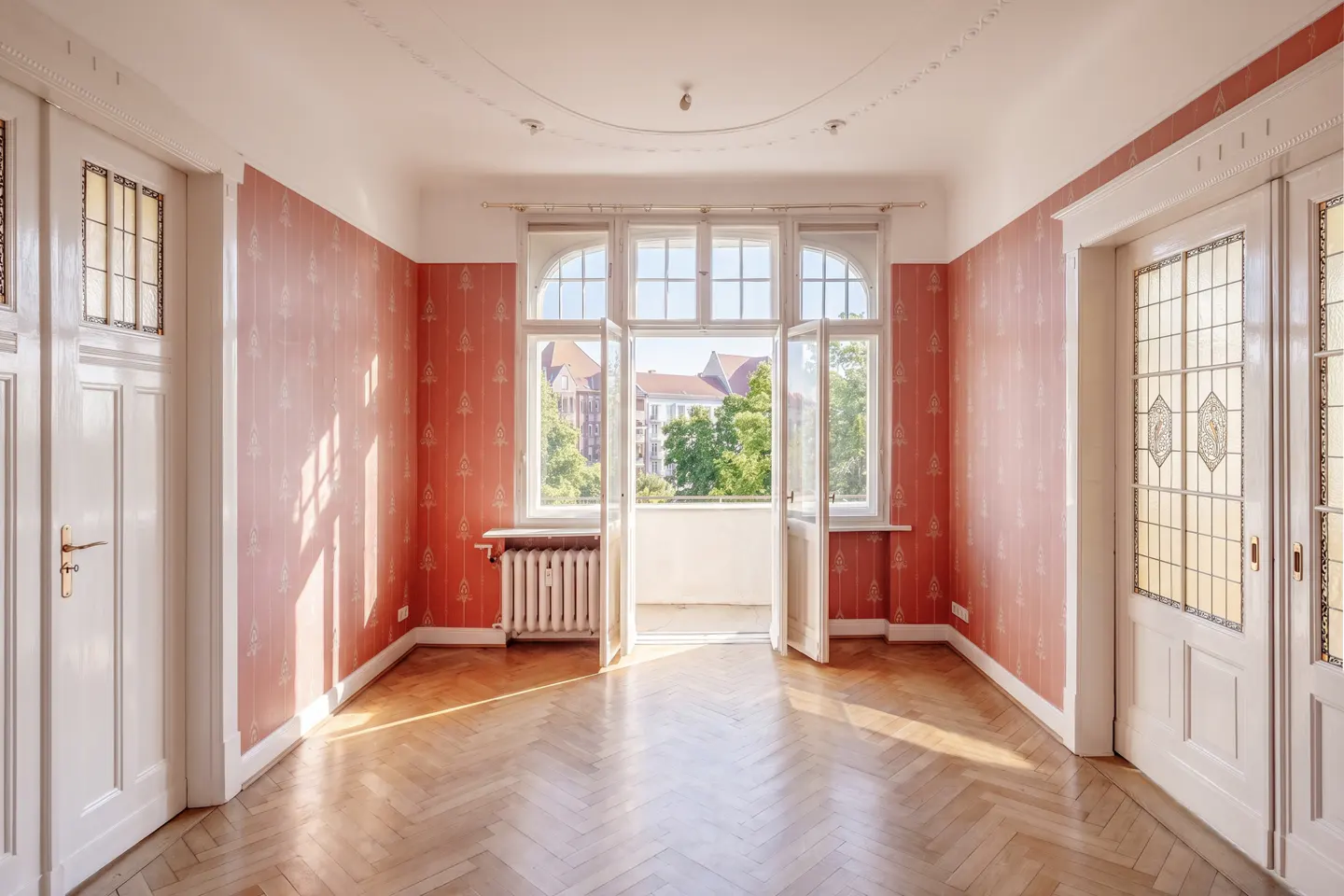 Empty room with herringbone wood floors, red patterned wallpaper, and white trim. Open doors lead to a balcony with a city view.