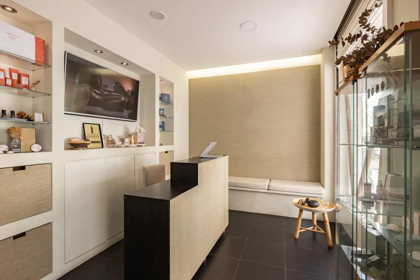Modern spa reception area with a black and beige desk, display shelves, and a waiting bench. A TV screen is mounted on the wall.