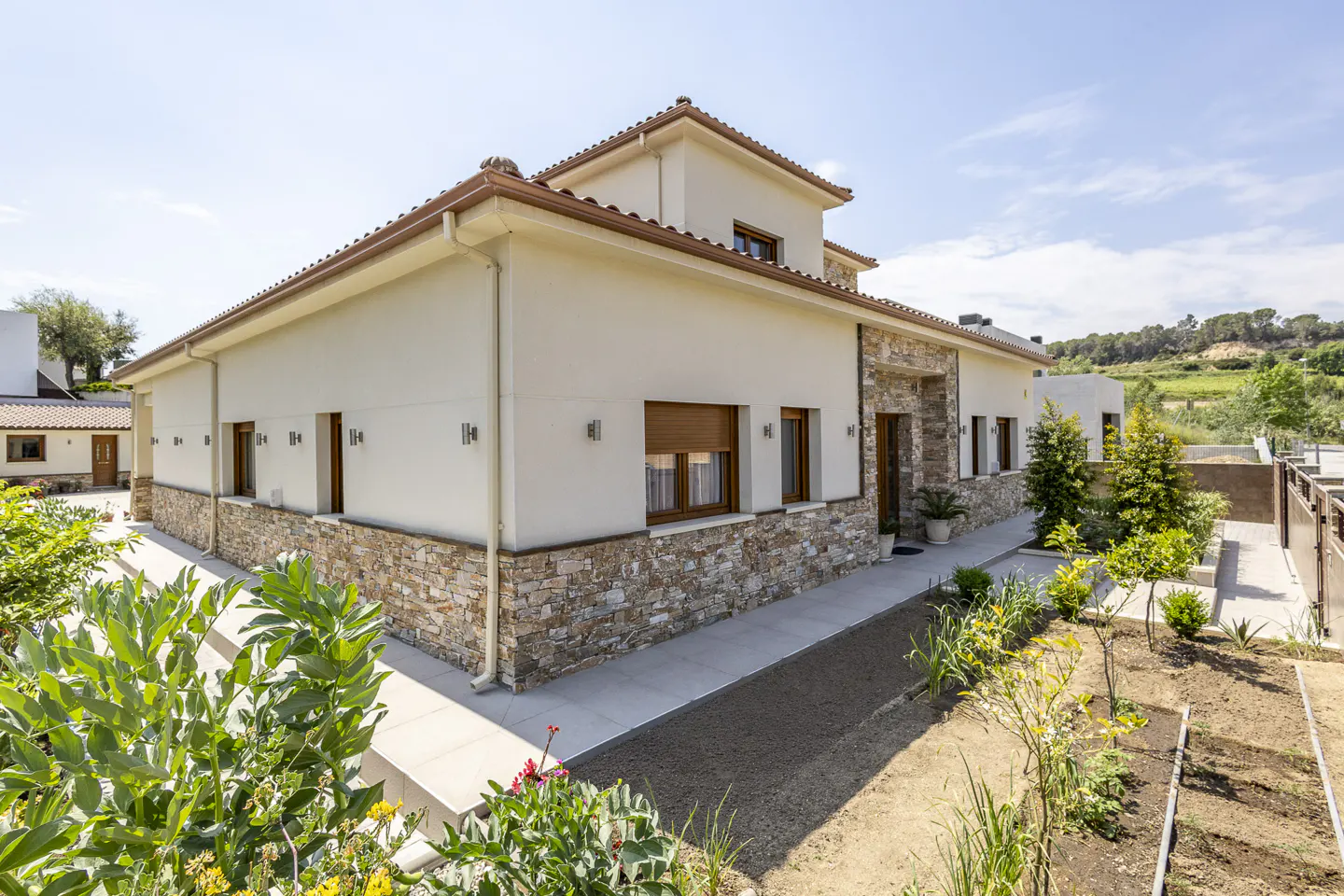 Beige two-story house with stone accents, brown trim, and a tile roof, surrounded by greenery and a blue sky.