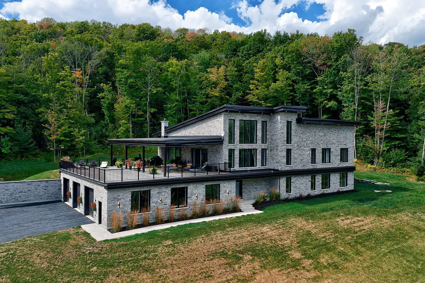 A modern, gray stone house with a black roof and trim sits on a green lawn with a forest backdrop.