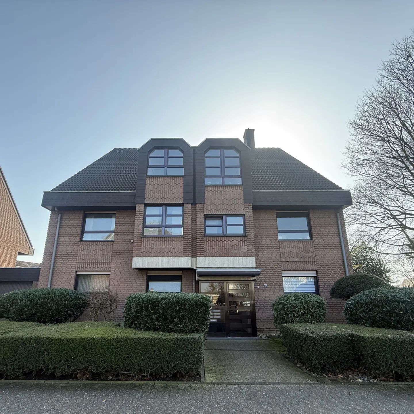 Brick apartment building with a dark roof and trimmed hedges in front.