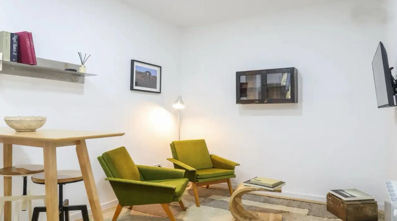 Living room with two green armchairs, a wooden table, and white walls. A shelf with books and a picture frame decorate the wall.