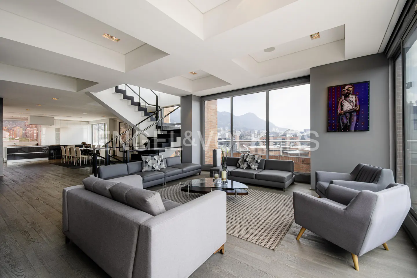 A modern living room with gray sofas, a glass table, and a staircase. Large windows offer a city view. An art piece hangs on the gray wall.