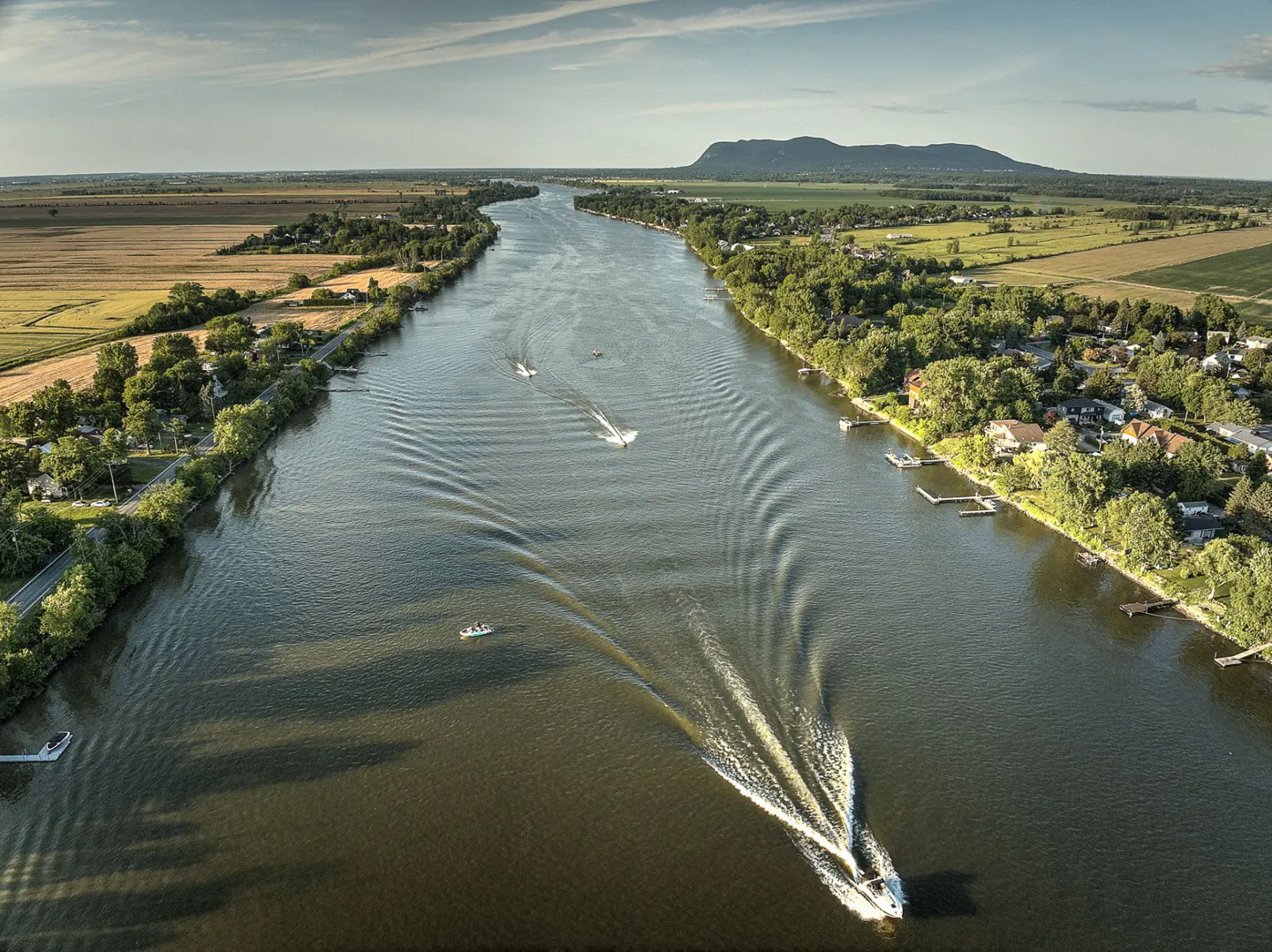 Aerial view of a wide river with boats leaving wakes, flanked by green trees and fields under a blue sky.