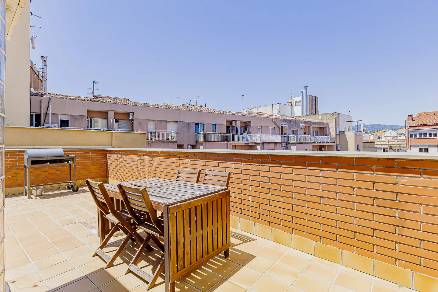 Outdoor patio with a wooden table, chairs, and grill. Brick wall surrounds the patio, with city buildings visible in the background.