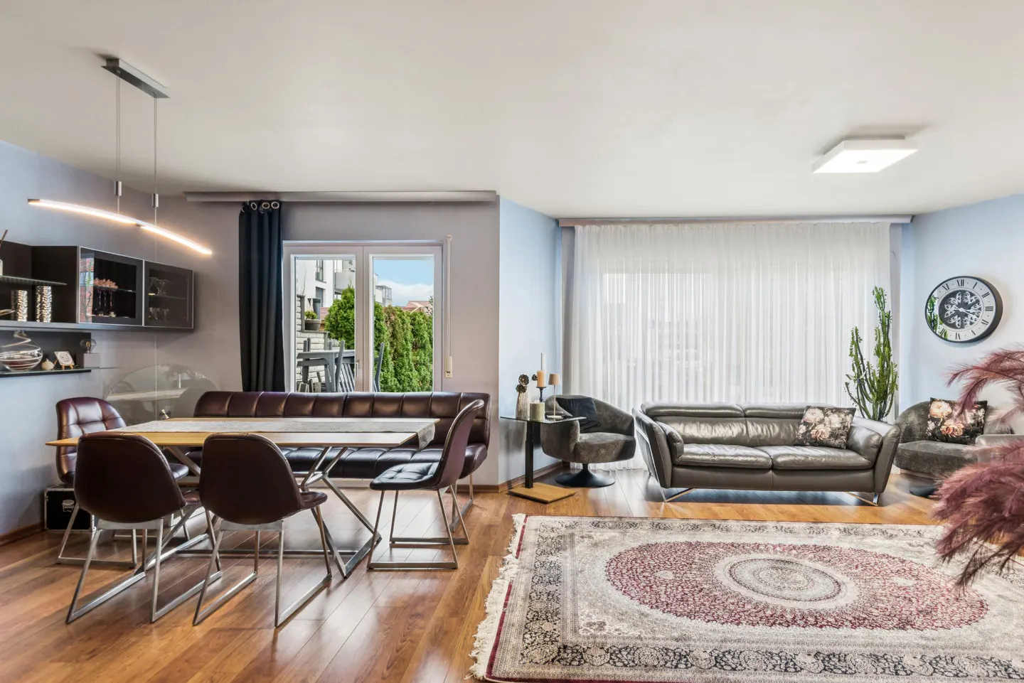 A modern living and dining area with a brown leather couch, dining bench, and chairs on a wood floor with a patterned rug.