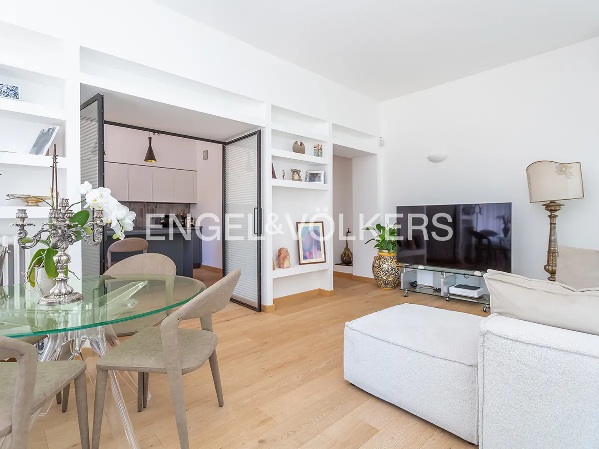 Bright, open-concept living space with a glass dining table, white sofa, and a view into the kitchen. Shelving units add storage.