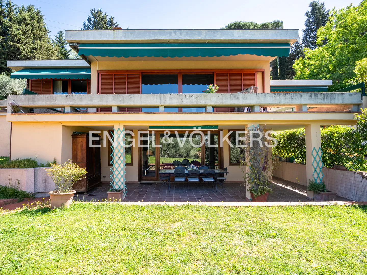 Two-story house with a green lawn. The house has a patio with a table and chairs. The Engel & Volkers logo is visible.
