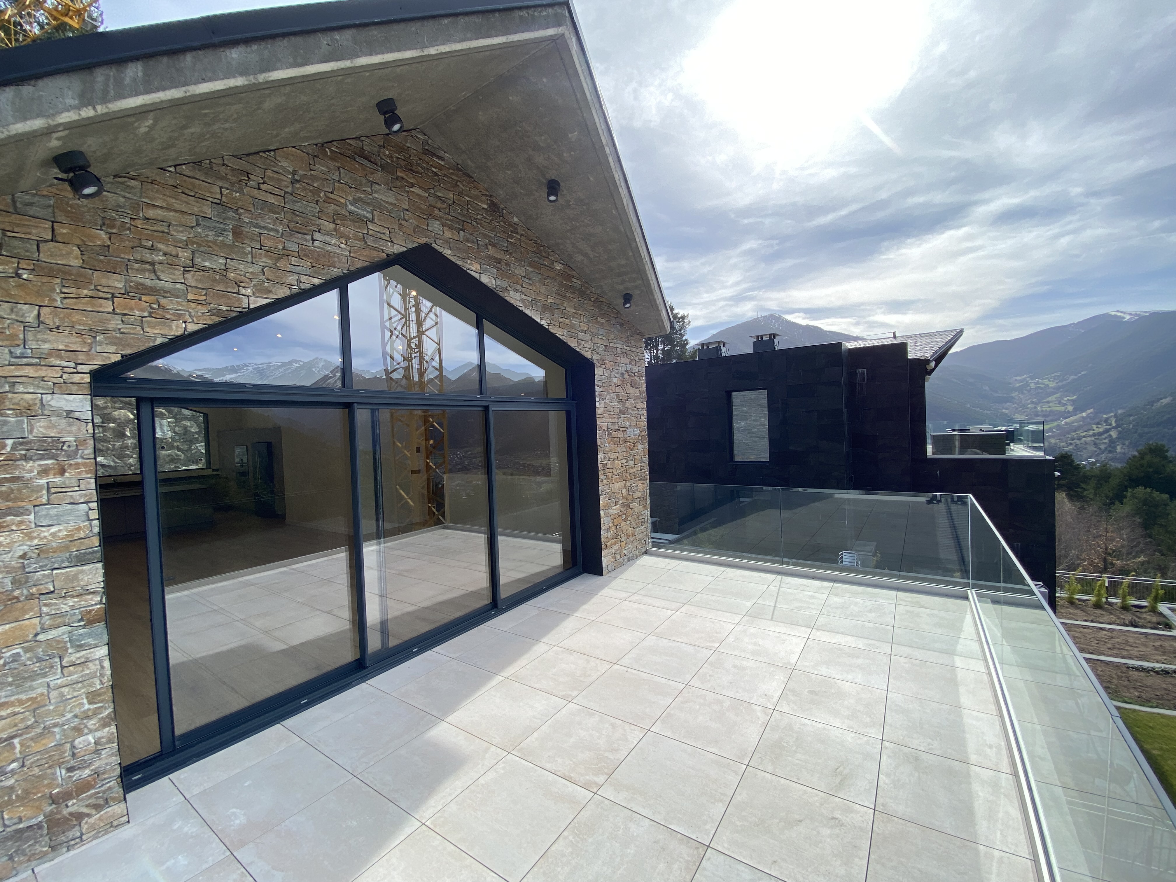 Exterior view of a modern house with stone walls, large windows, and a tiled patio with glass railings overlooking mountains.