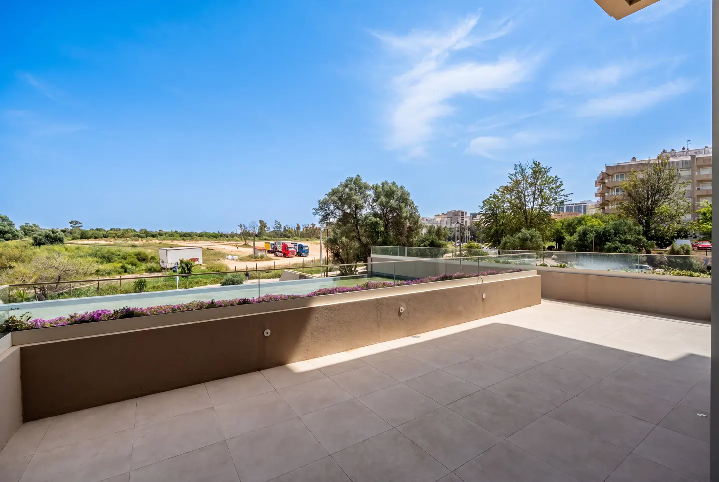 Wide shot of a beige tiled balcony with a low wall and glass railing overlooking a field and blue sky.