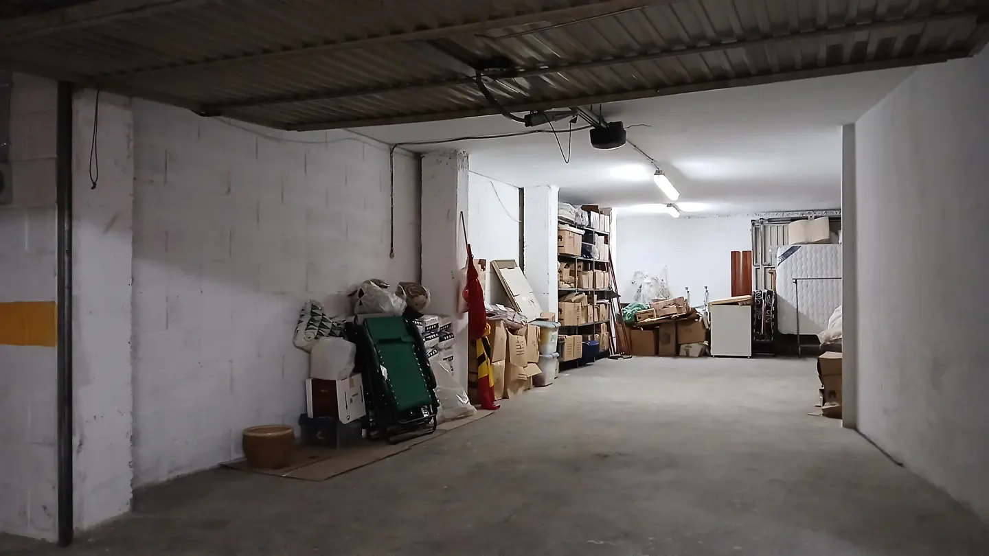 A spacious, dimly lit garage with white concrete walls, filled with storage boxes, a folding chair, and miscellaneous items.