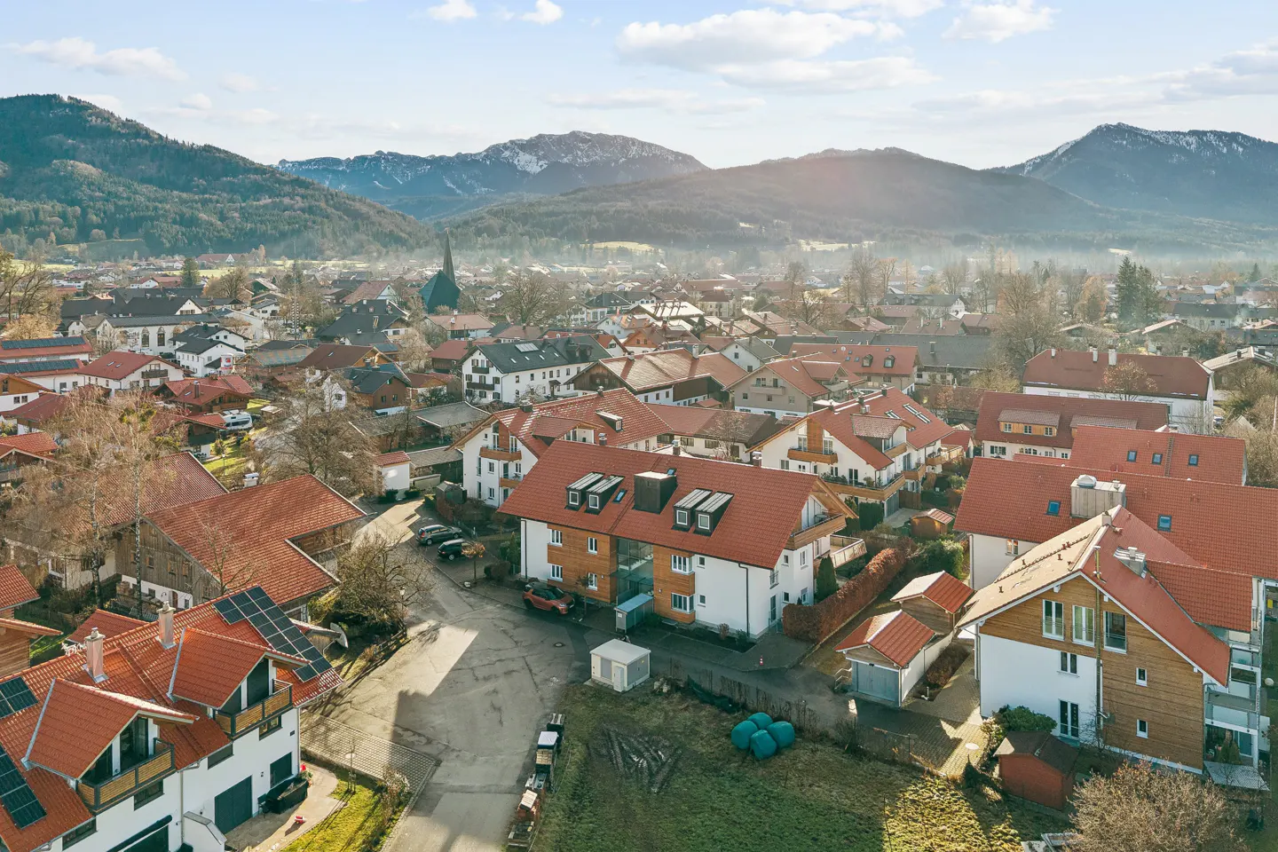 Aerial view of a European town with red-tiled roofs, a church steeple, and mountains in the background on a sunny day.