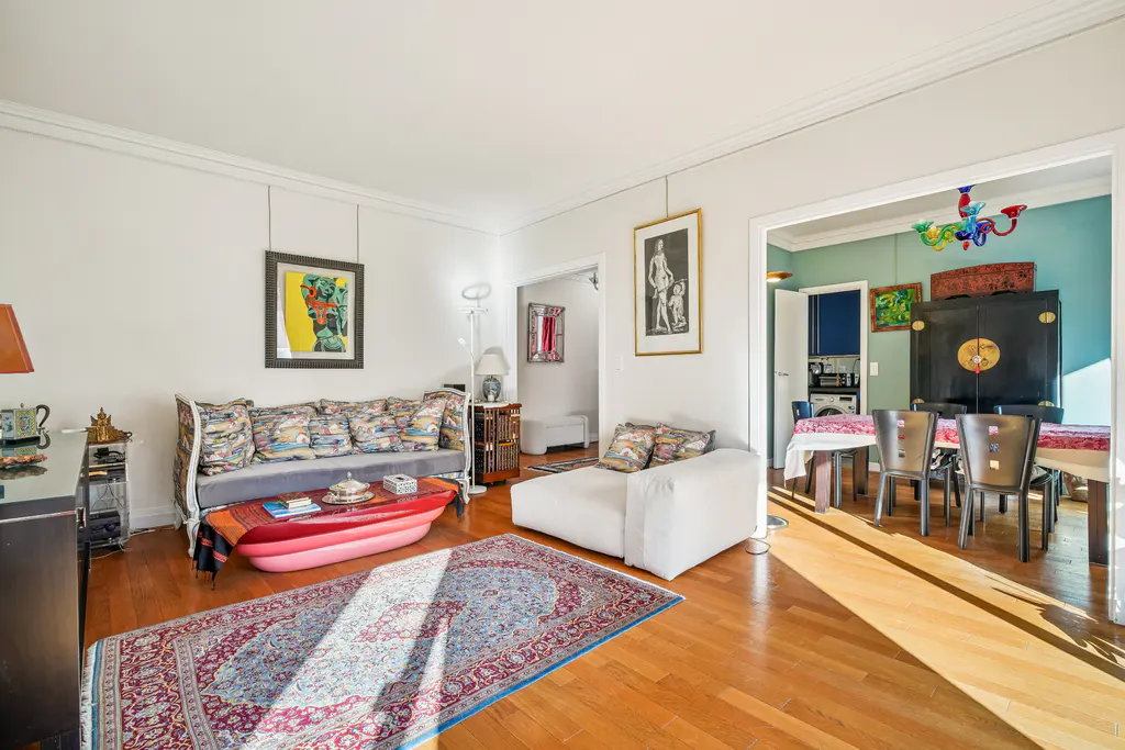 Bright living room with hardwood floors, patterned rug, sofa, chaise lounge, and artwork. Dining room visible through doorway.