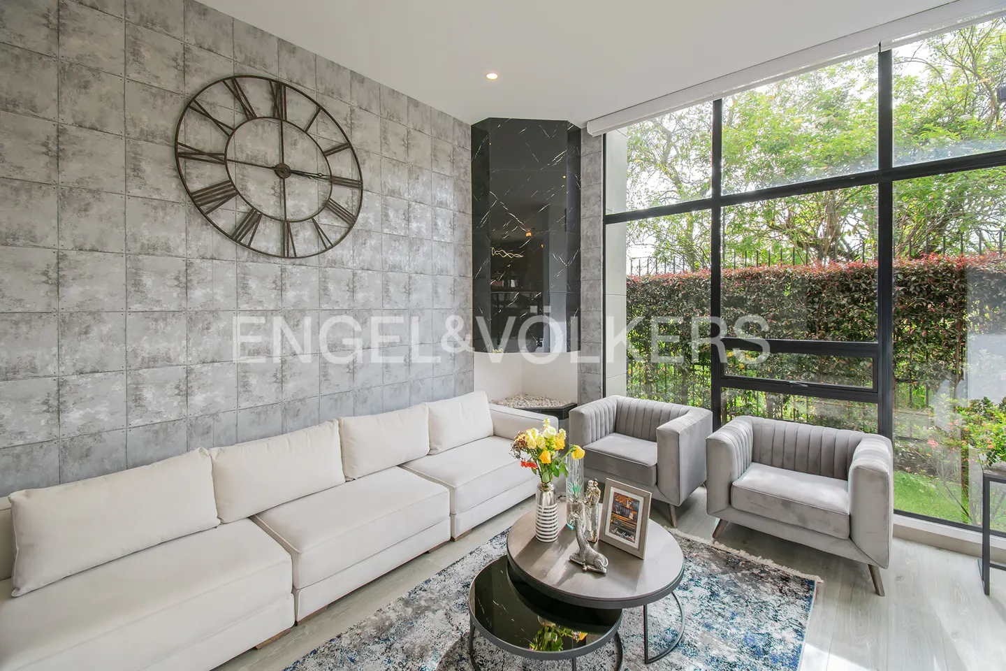 Living room with white sofa, gray chairs, and a large window overlooking greenery. A clock hangs on the gray tiled wall.