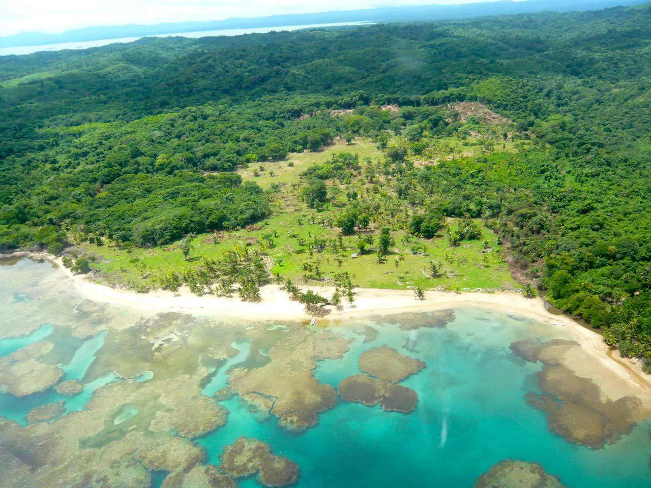 Aerial view of a tropical beach with turquoise water, coral reefs, white sand, and lush green forest.