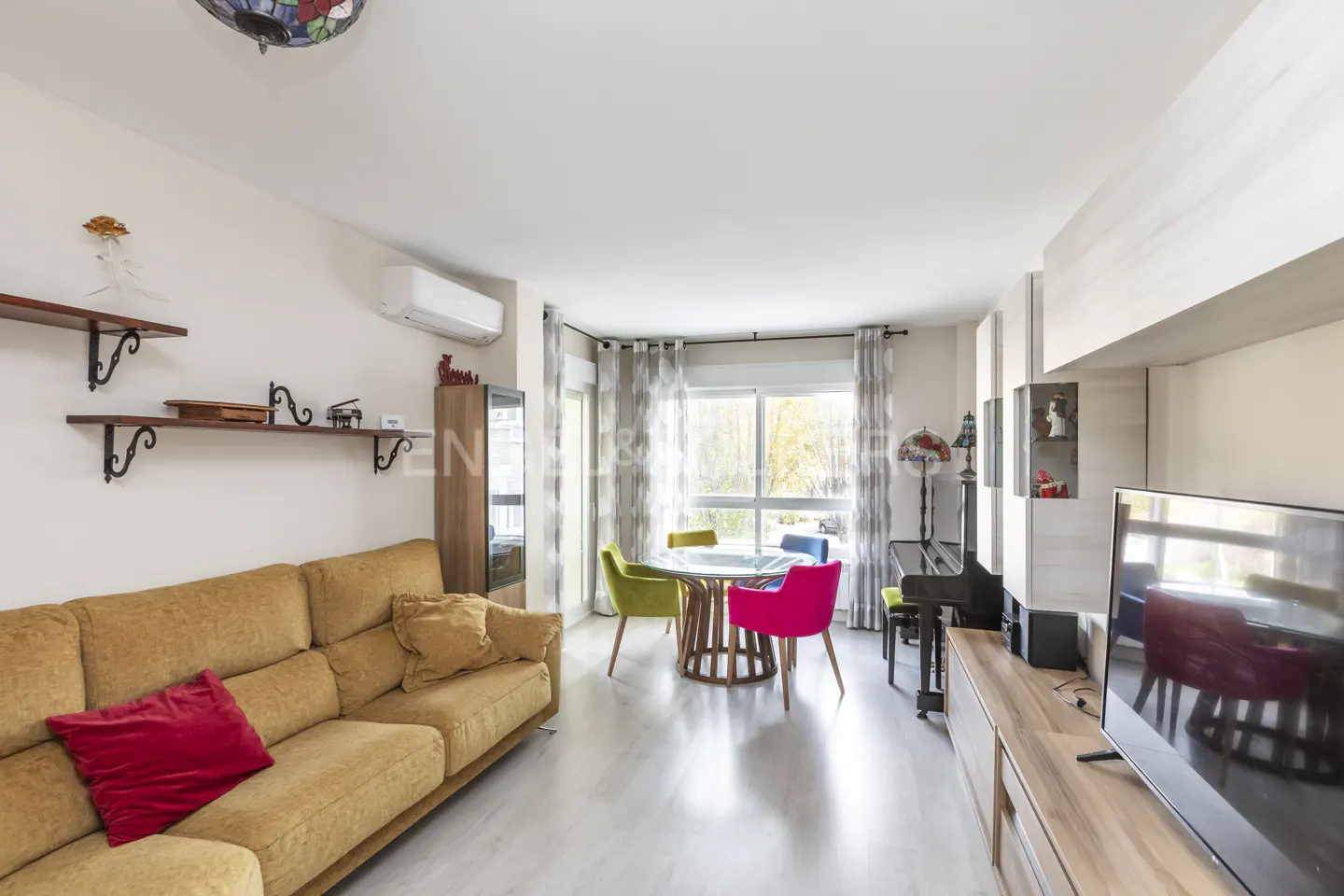 Bright living room with a yellow sofa, red pillow, round table, colorful chairs, piano, and a large TV.