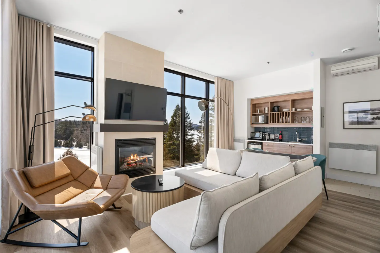 Bright living room with a fireplace, TV, and large windows overlooking a snowy landscape. A tan leather chair and white sofa provide seating.