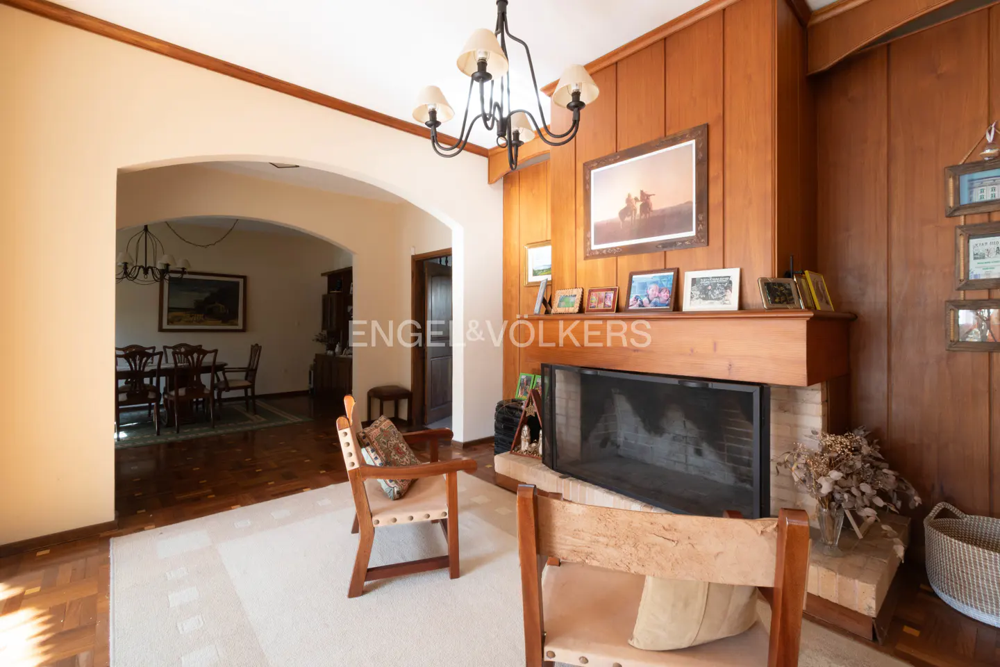 Living room with wood paneled walls, fireplace, and two chairs. A dining room is visible through an arched doorway.