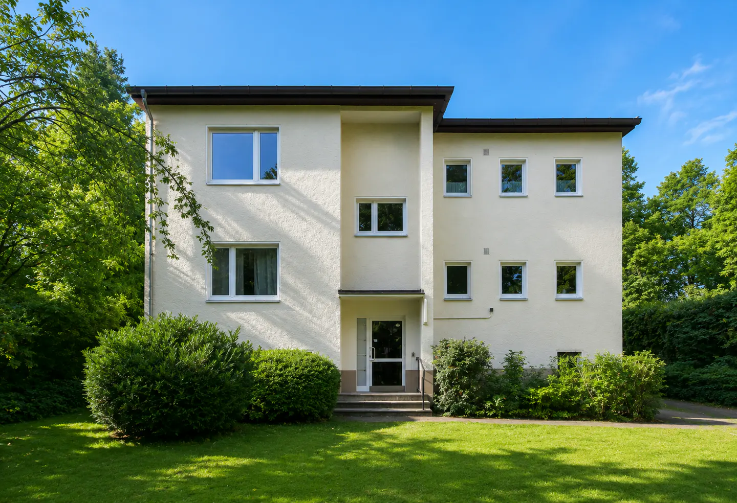 A three-story cream-colored apartment building with a flat roof, surrounded by lush green trees and a manicured lawn.