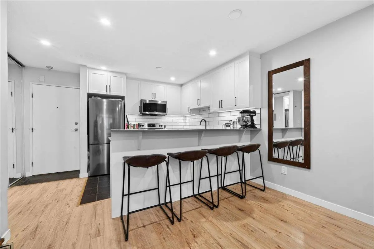 Bright kitchen with white cabinets, stainless steel appliances, and a breakfast bar with four brown leather stools. A wood-framed mirror hangs on the wall.