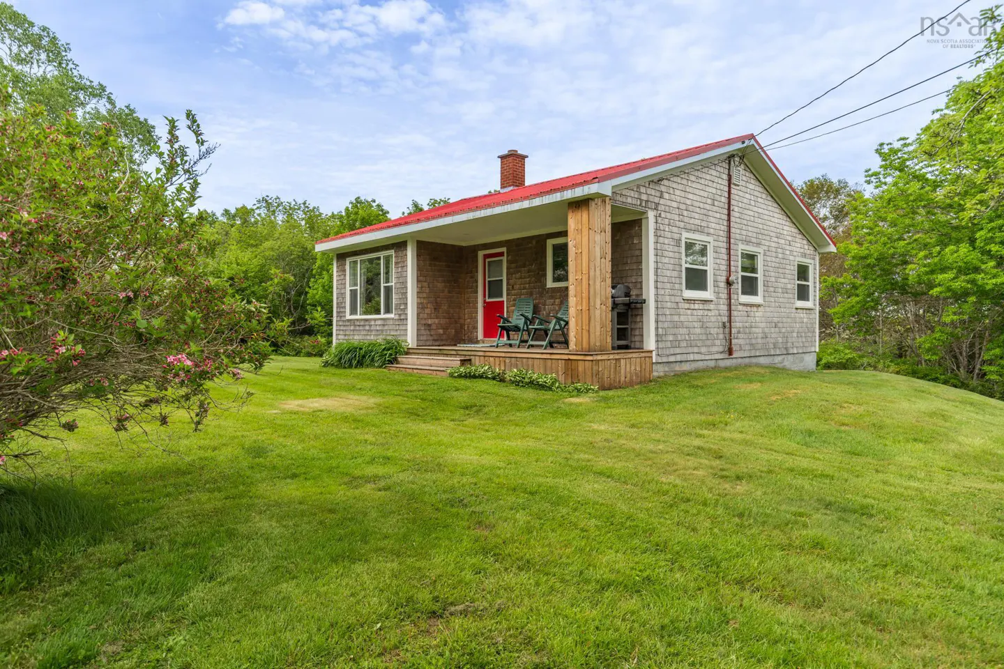 A cottage with grey shingle siding, a red roof, and a red front door, set on a grassy lawn with trees in the background.