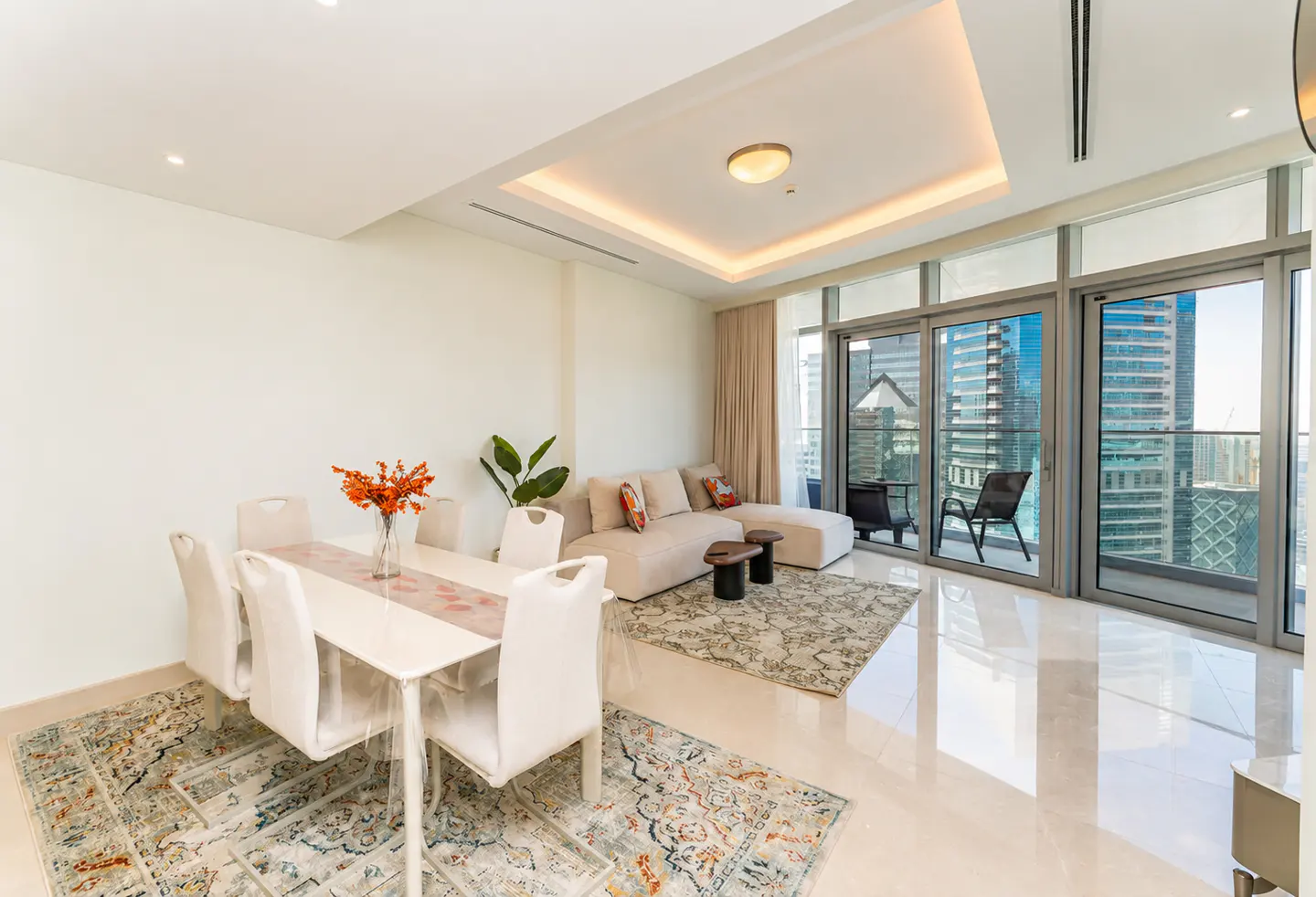 Bright, modern apartment interior with a dining table, sofa, and balcony overlooking a city skyline. Cream walls and tile floors.