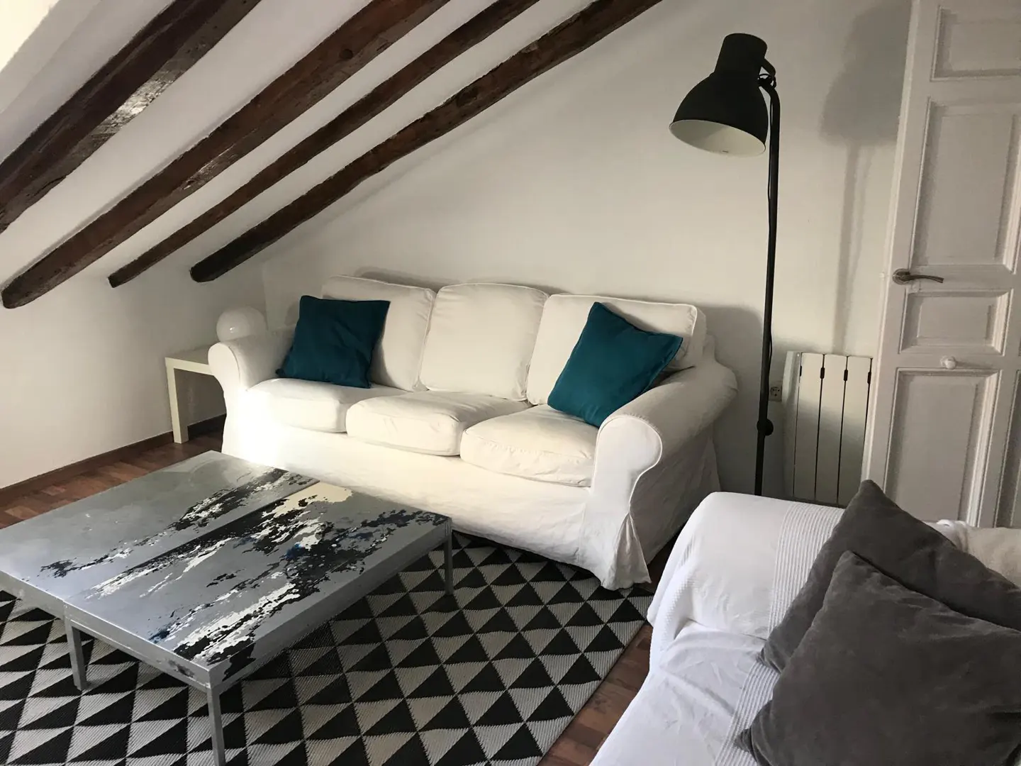 Attic living room with white sofa, teal pillows, black floor lamp, and a patterned rug under a modern coffee table.