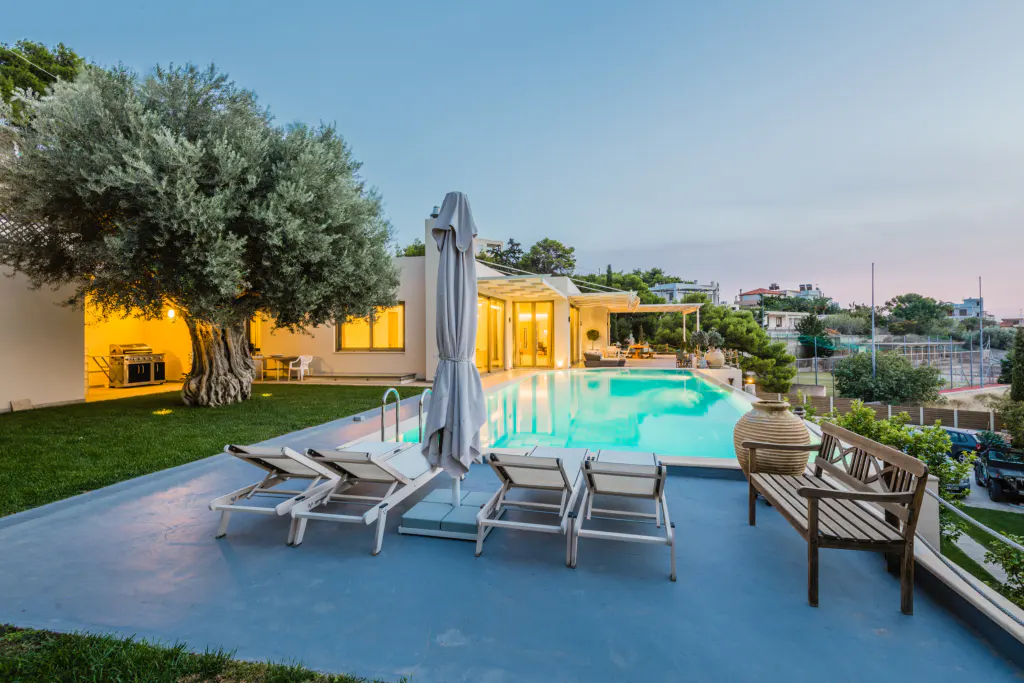 Outdoor view of a luxury home with a pool, lounge chairs, a bench, and a large tree at dusk.