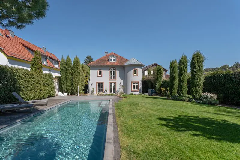 Exterior view of a gray two-story house with a pool, green lawn, and tall trees under a clear blue sky.