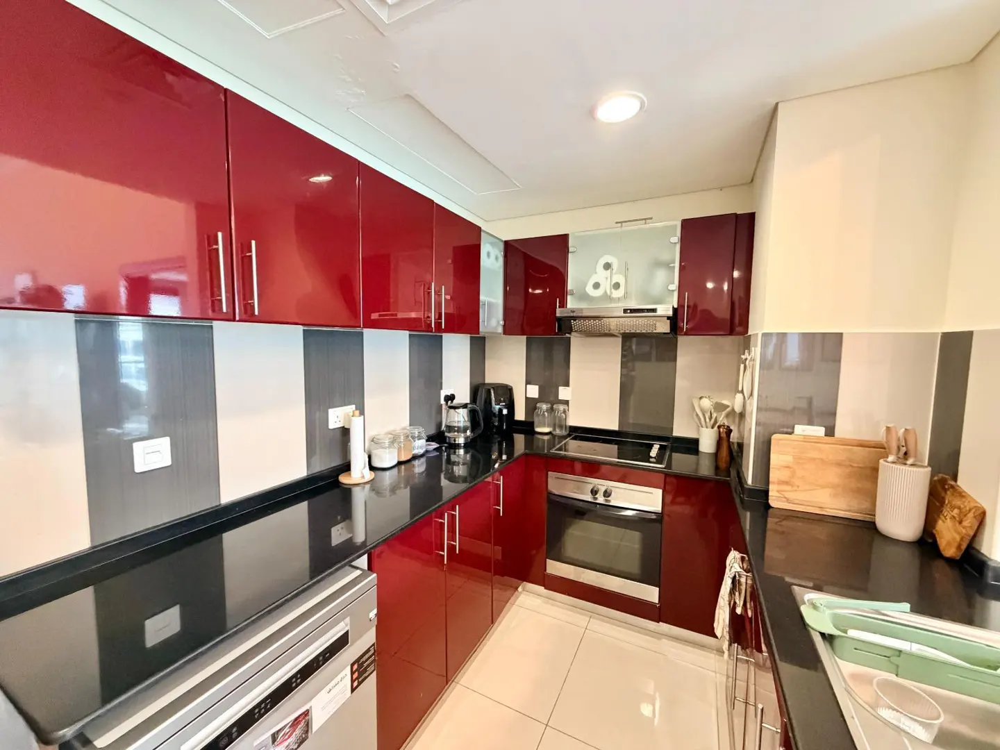 A modern kitchen with red cabinets, black countertops, and striped gray and white backsplash tiles. Appliances are visible.
