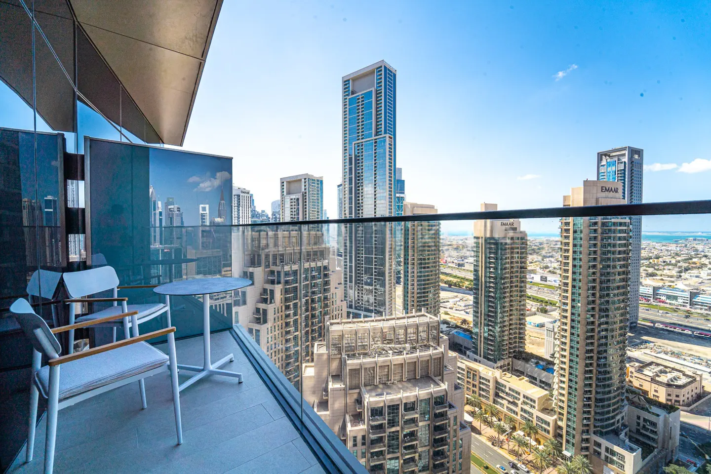 Balcony view of Dubai skyline. Two white chairs and a table sit on the balcony overlooking tall buildings under a blue sky.