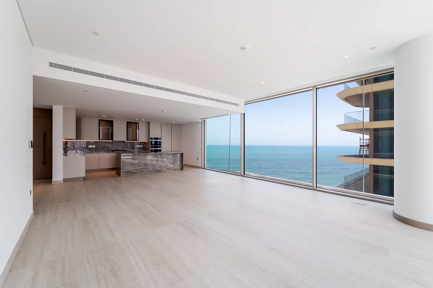 A bright, modern apartment interior with a kitchen, marble island, and floor-to-ceiling windows overlooking the ocean.