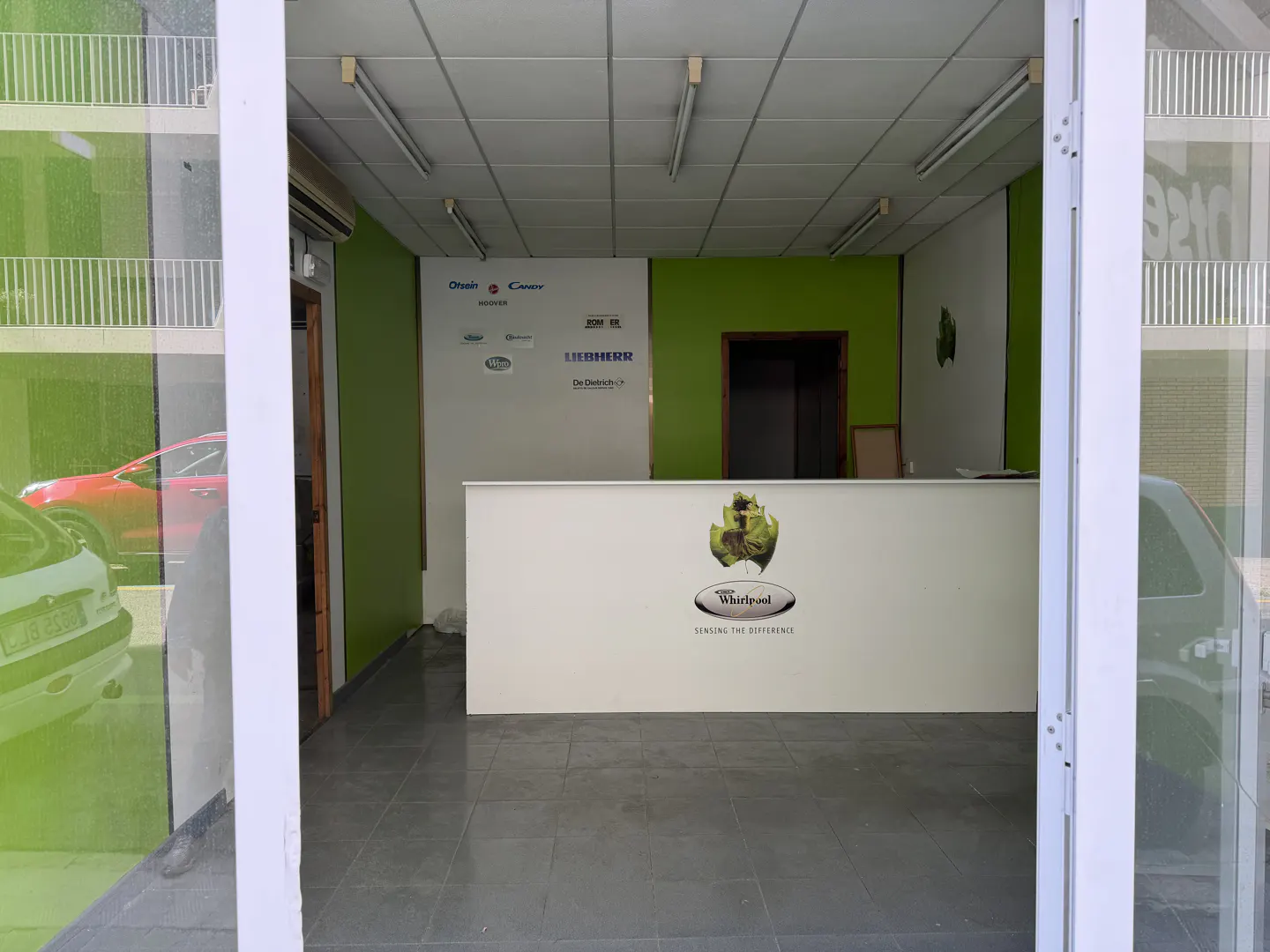 View through open white doors into a commercial space with a white reception desk featuring the Whirlpool logo. Green accent walls and appliance brand logos are visible.