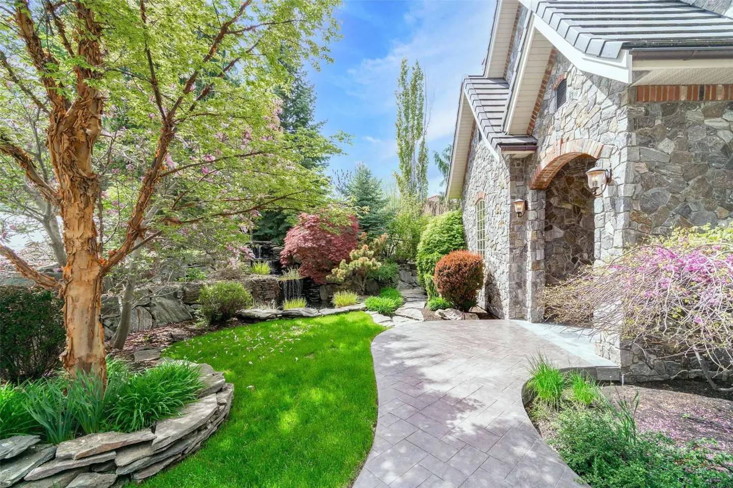 Stone house exterior with a curved walkway, green lawn, trees, and a small waterfall feature in the background.