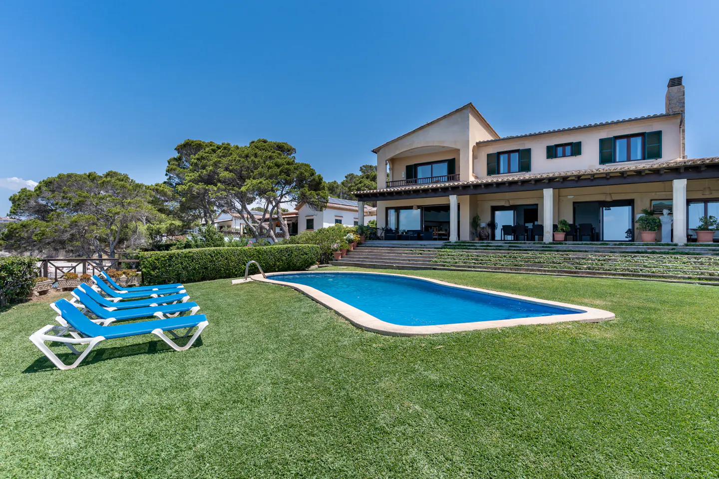 A tan two-story house with a pool and blue lounge chairs on a green lawn under a blue sky.