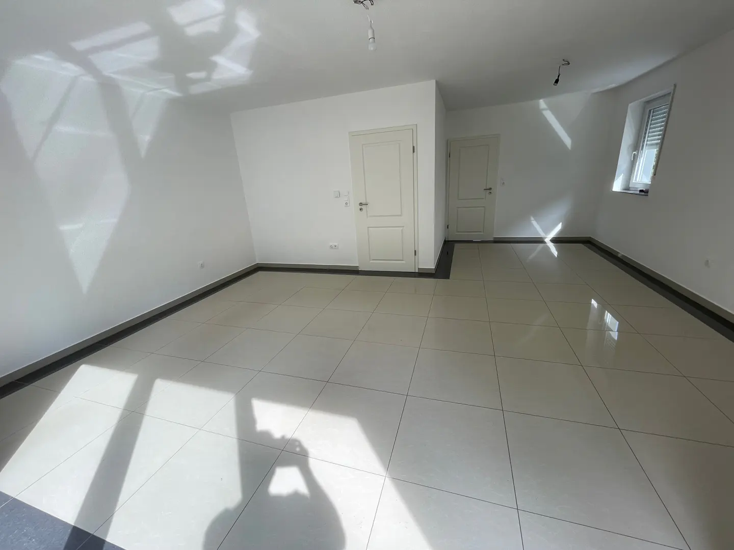 Bright, empty room with white walls, tile floor, and two white doors. Sunlight streams in from a window with closed blinds.