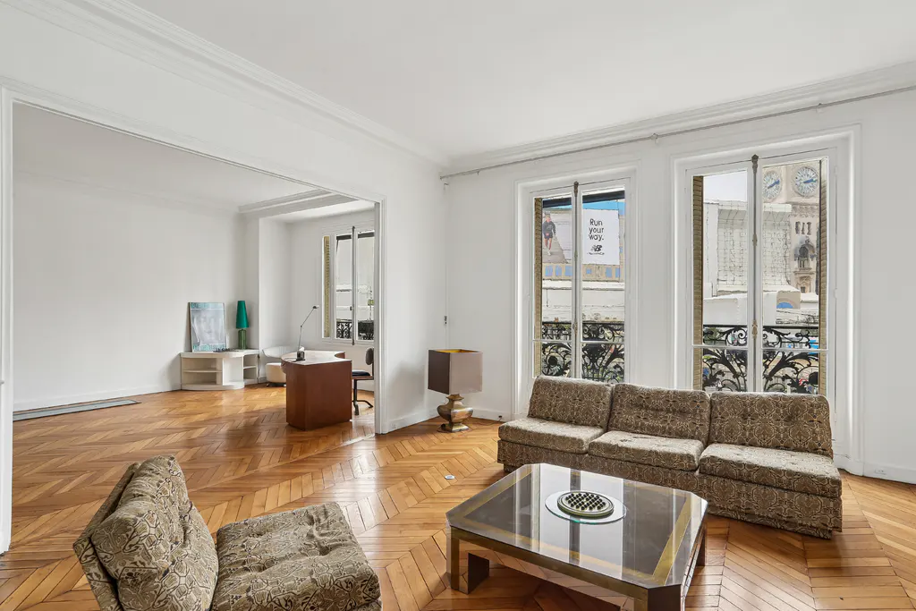 Bright living room with herringbone wood floors, patterned sofa and chair, and a view of a clock tower through the windows.