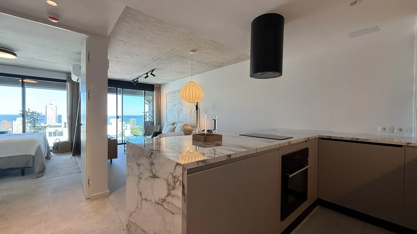 Open-concept apartment with marble kitchen island, black range hood, and ocean view from the living and bedroom areas.