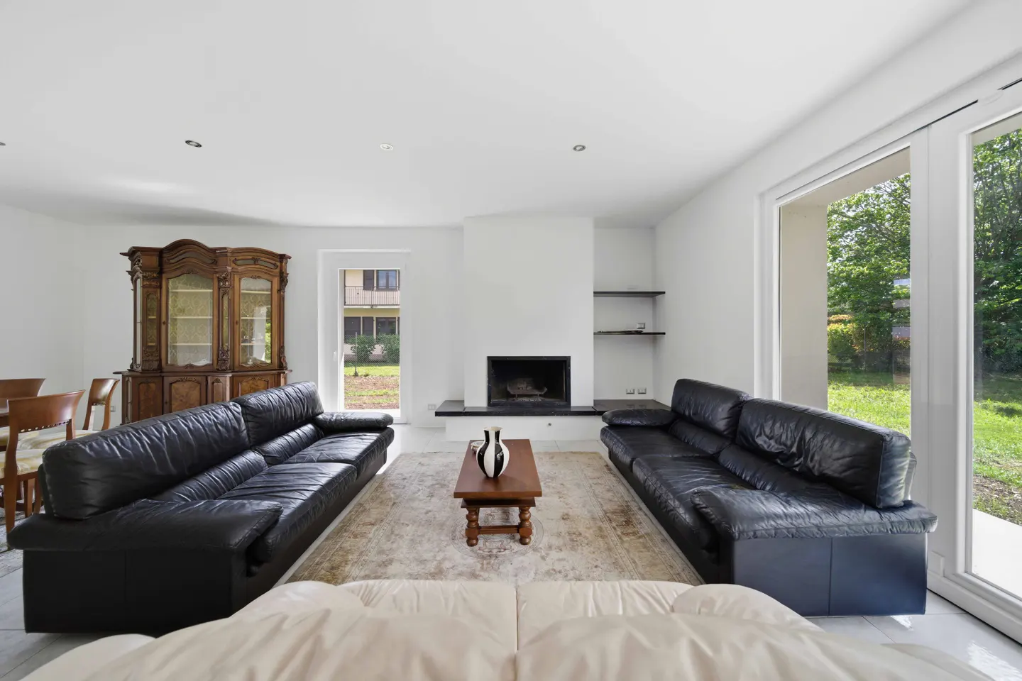 Bright living room with black leather sofas, a fireplace, and a wooden cabinet. A beige rug covers the floor.