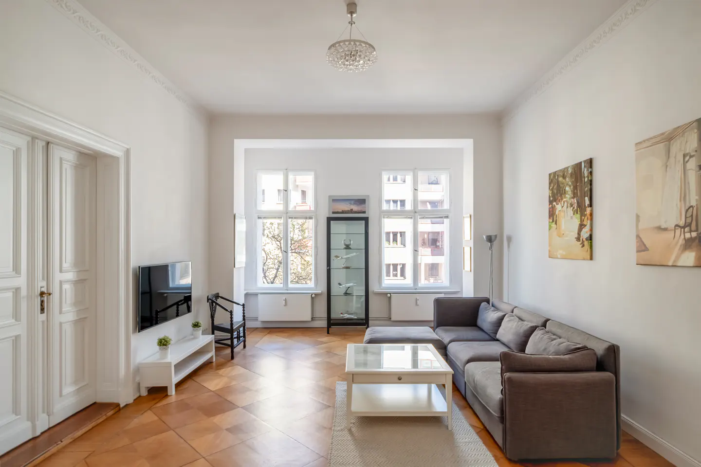 Bright living room with hardwood floors, white walls, and large windows. A gray sectional sofa faces a TV. Artwork adorns the walls.