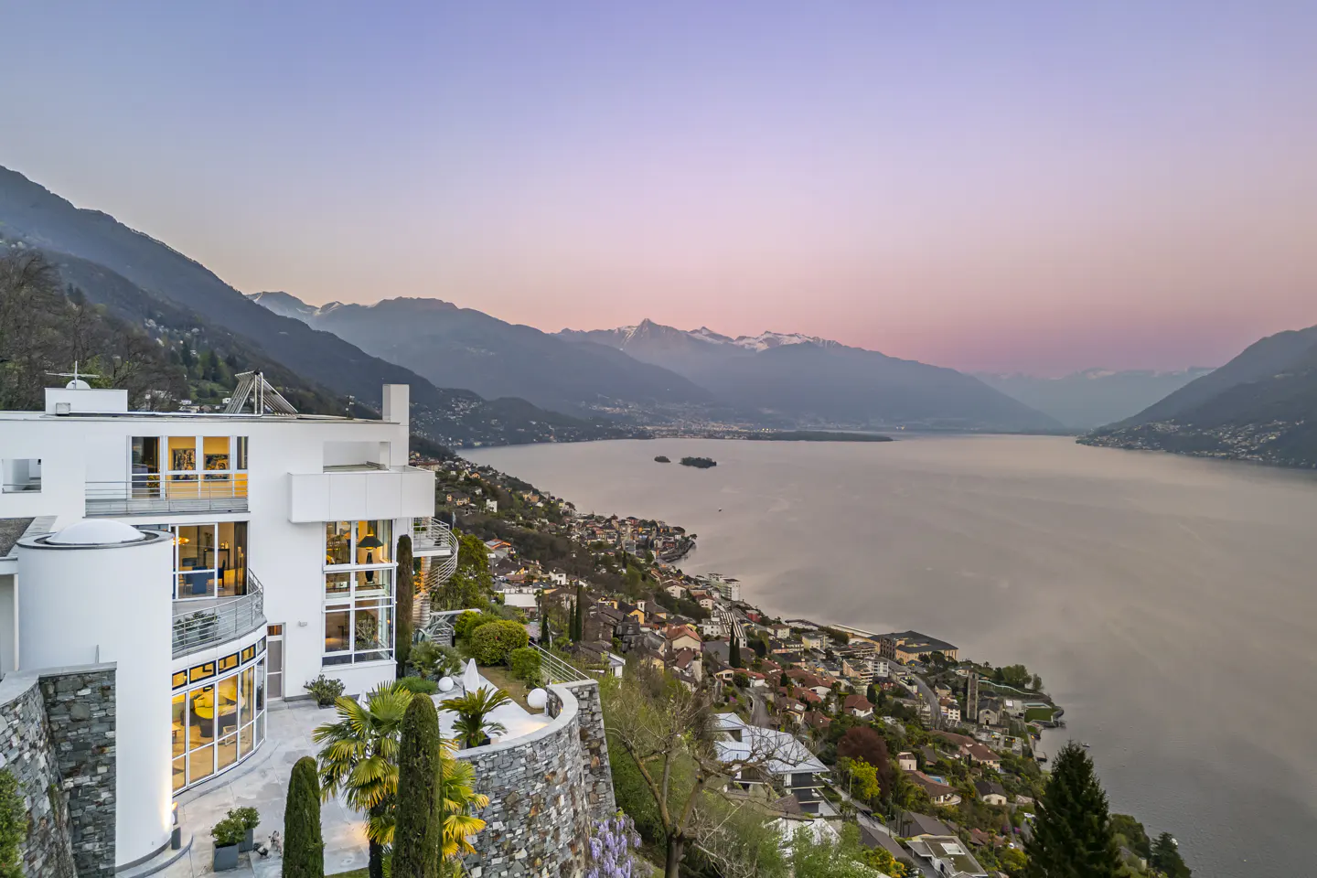 Modern white house with large windows overlooking a lake and mountains at sunset.