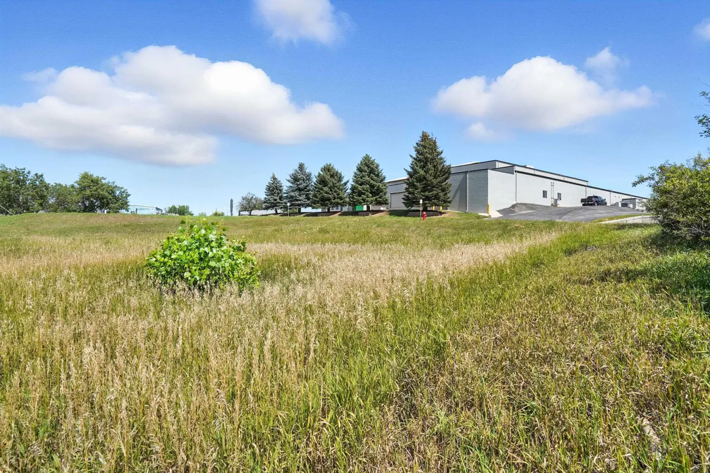 Exterior shot of a gray commercial building on a grassy hill under a blue, cloudy sky. Tall grass in the foreground.