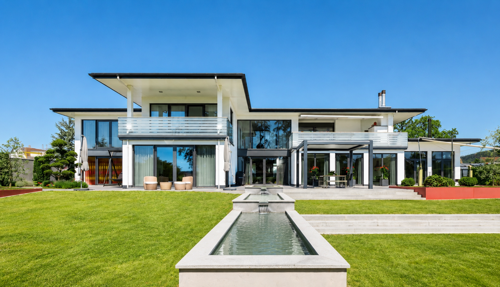 Modern two-story white home with large glass windows, a tiered water feature, and a lush green lawn under a clear blue sky. [AI-generated text]