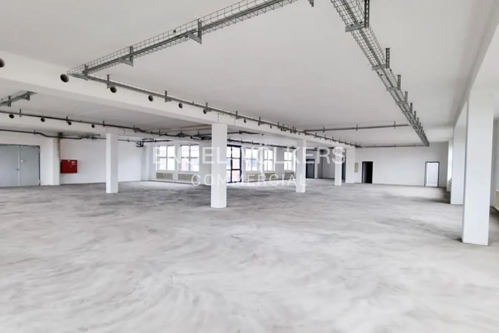 Bright, empty commercial space with white walls, concrete floors, and support columns. Metal cable trays run along the ceiling.