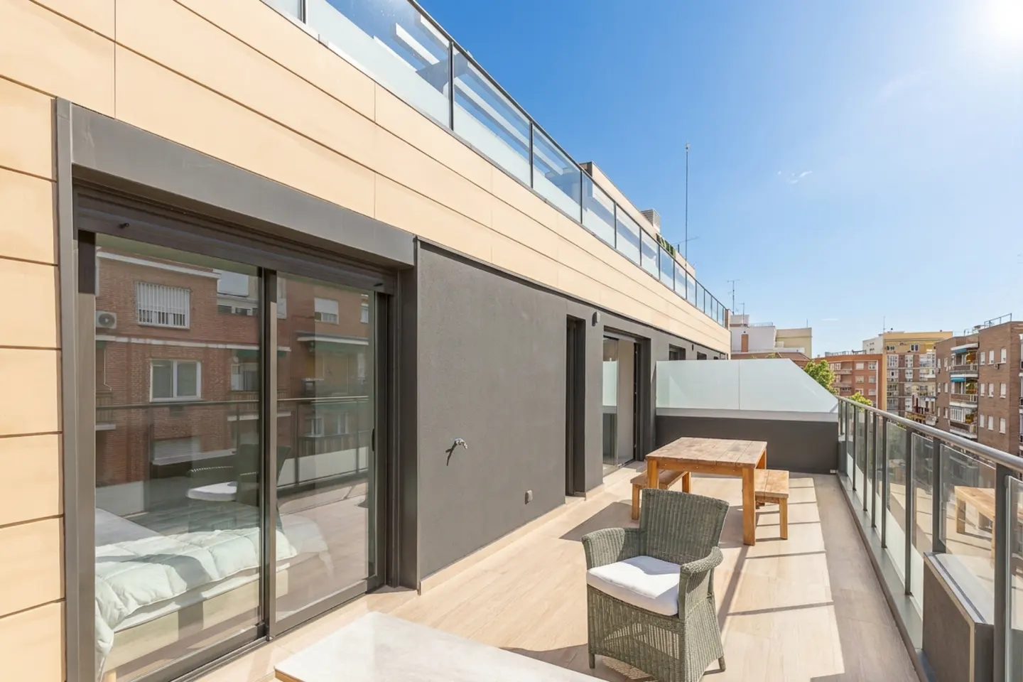 A sunny balcony with a wicker chair, wooden table, and glass railings overlooks a cityscape under a clear blue sky.