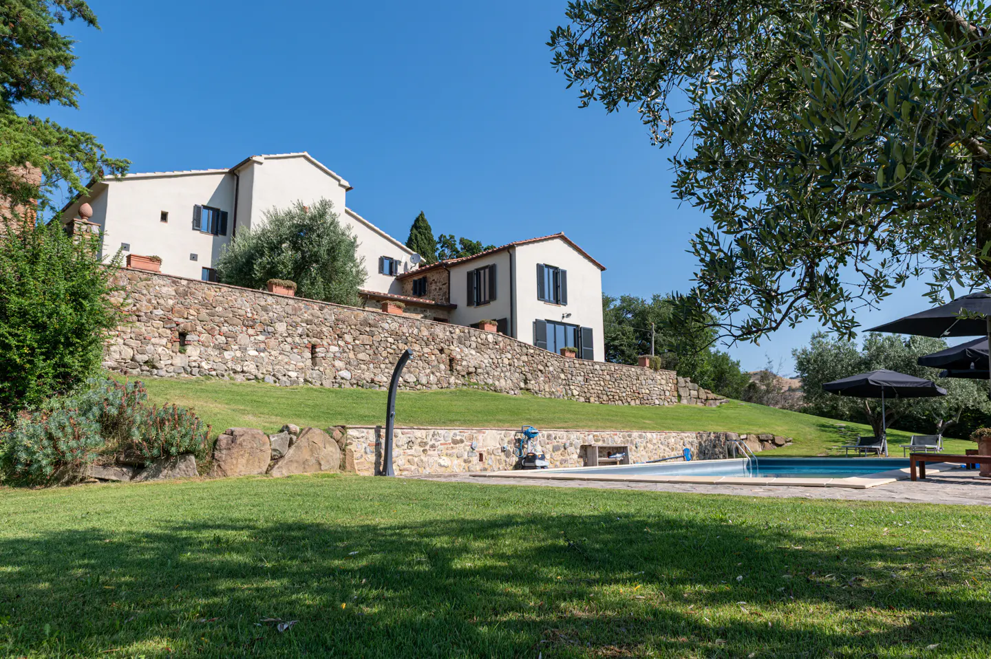 Exterior view of a two-story white house with black shutters, a stone wall, a pool, and green grass.