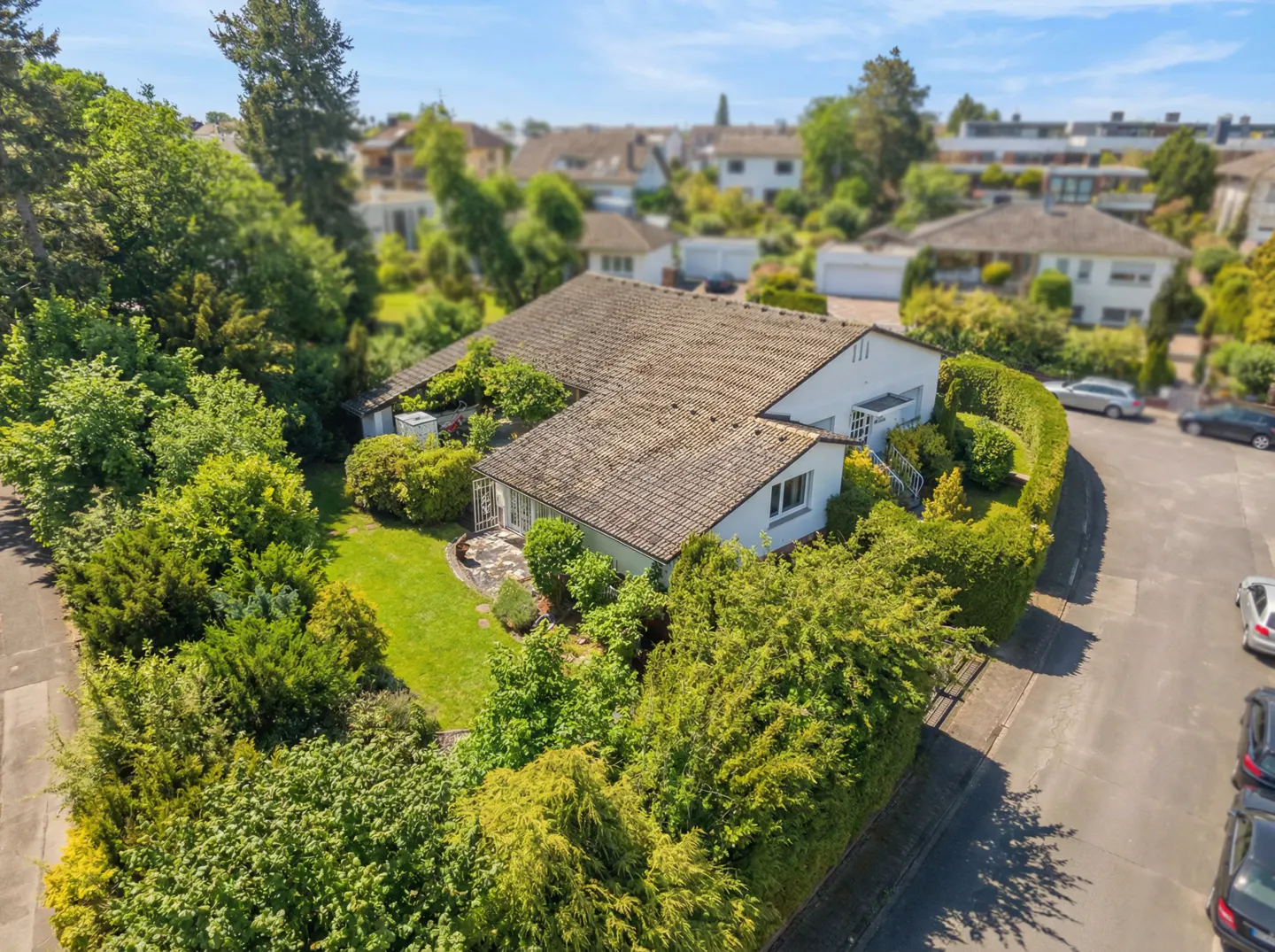Aerial view of a white, single-story house with a brown tile roof, surrounded by lush green trees and bushes. Cars are parked on the street.