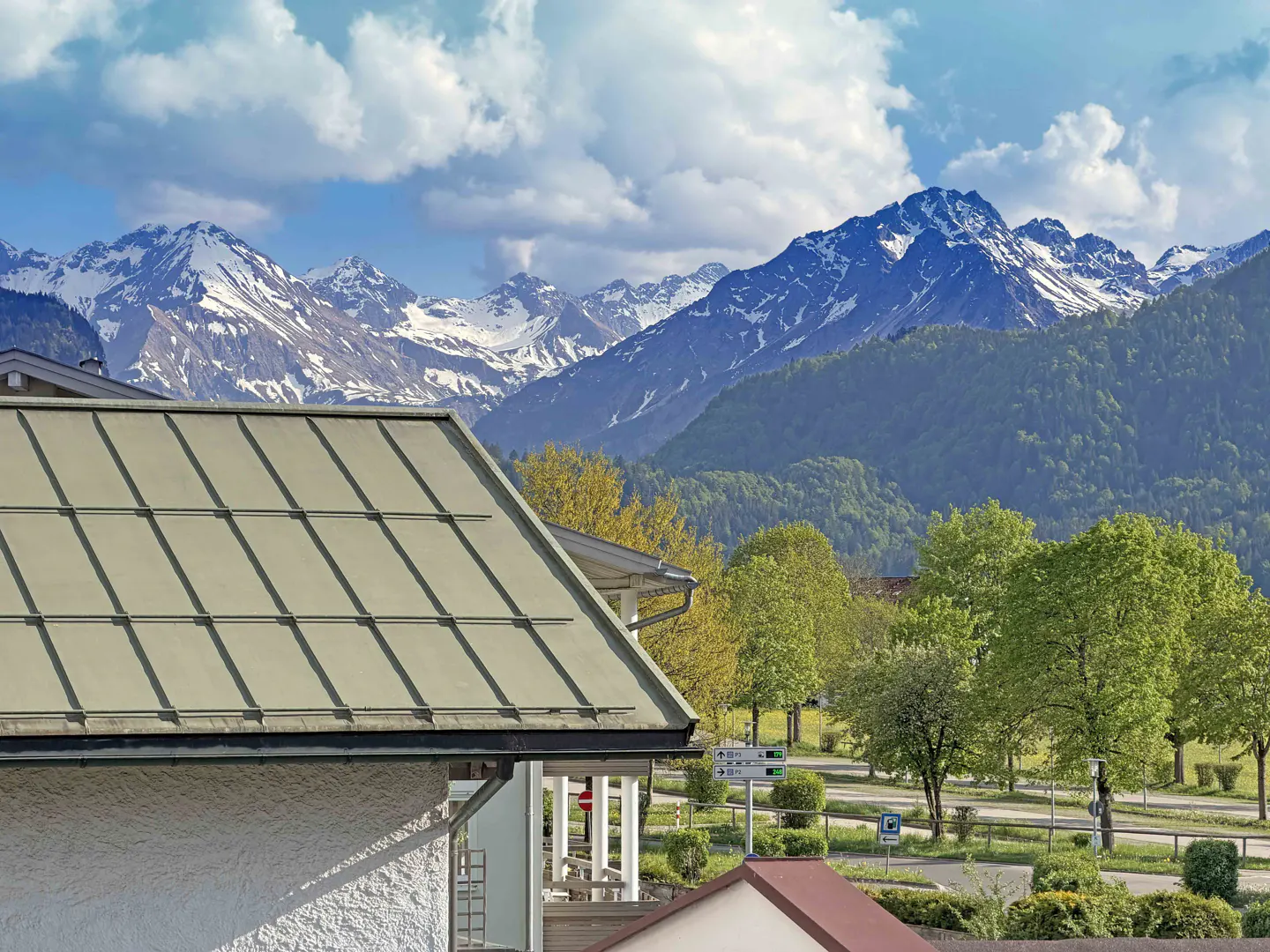 View of snow-capped mountains behind a building with a green roof and a parking lot with trees.